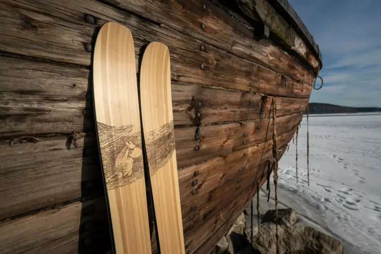 Ein Paar PUSU Moomin Skier lehnt an der rustikalen, dunklen Holzwand eines alten Bootes an einem gefrorenen See. Die natürliche Holzmaserung der Skier kontrastiert mit der detaillierten Mumin-Grafik im oberen Bereich.