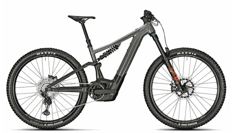 Premium E-MTB auf höchstem Niveau 7 Ein vollgefedertes E-MTB in Titansilber mit einem markanten Stahlfederdämpfer im Rahmen, Maxxis-Reifen und orangefarbenen Fox-Gabel-Decals.