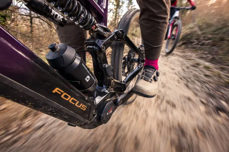 Eine dynamische Nahaufnahme aus der Froschperspektive während der Fahrt auf einem E-Mountainbike von Focus. Im Fokus stehen der Rahmen mit dem Bosch-Motor, ein Fox-Stahlfederdämpfer, eine Trinkflasche und der Fuß des Fahrers in einem beigefarbenen Klickschuh mit pinker Socke auf dem Pedal. Der staubige Trail unter den Reifen ist durch die Geschwindigkeit verwischt.