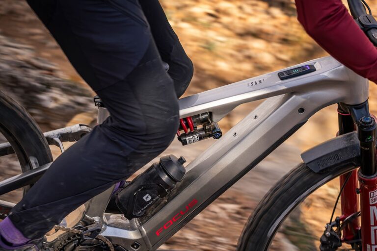 Seitenansicht des silbernen Aluminiumrahmens eines Focus SAM² E-Mountainbikes während der Fahrt. Deutlich zu sehen sind das im Oberrohr integrierte Display mit LED-Akkuanzeige, der rote RockShox-Stahlfederdämpfer im Hinterbau und eine Trinkflasche im Unterrohr. Der Fahrer trägt eine schwarze Hose, während die rote RockShox-Federgabel das Vorderrad führt.
