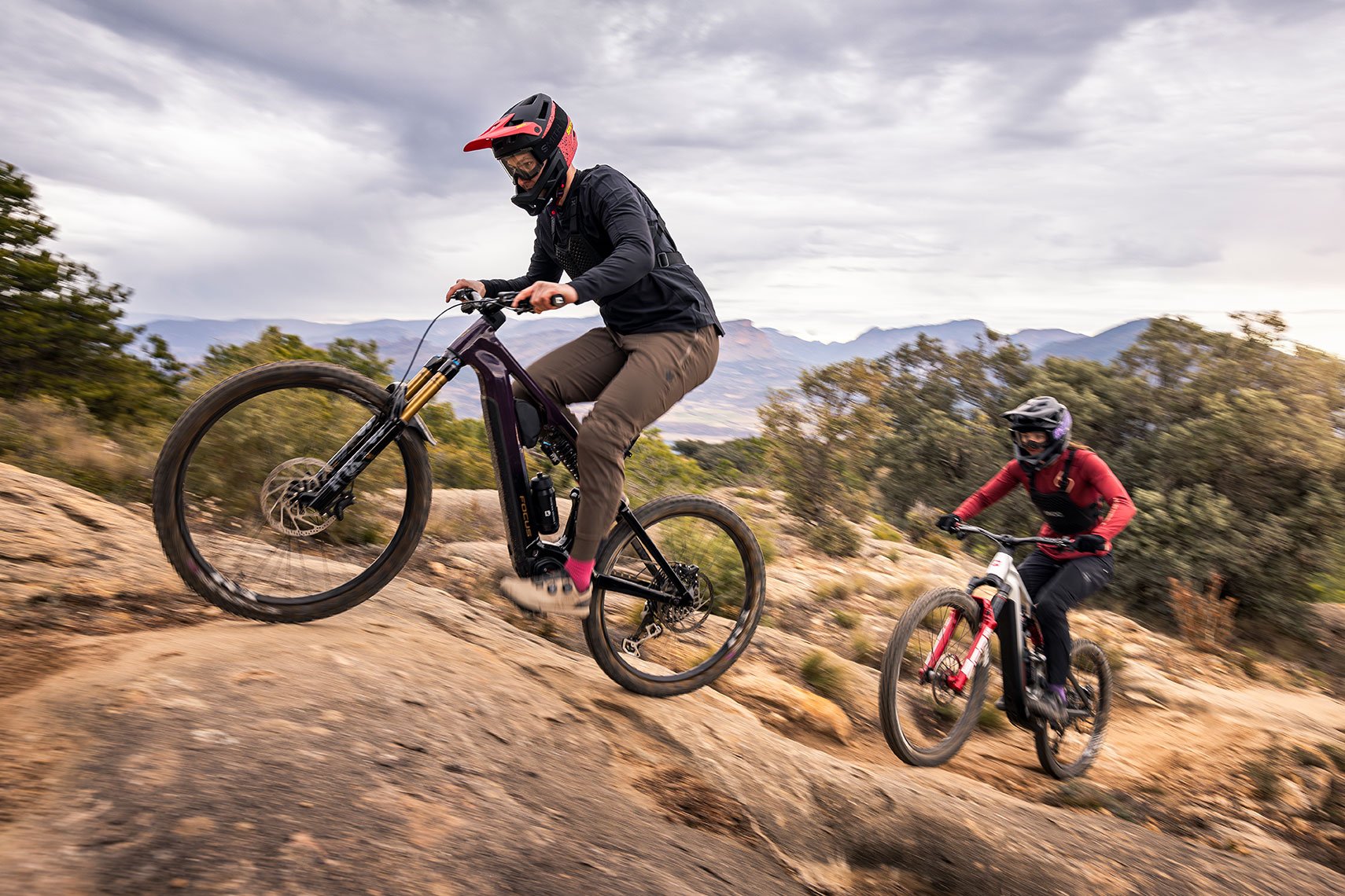 Zwei Mountainbiker in voller Schutzkleidung und mit Integralhelmen fahren auf einer felsigen, steilen Bergab-Passage. Im Vordergrund fährt eine Person auf einem dunkelvioletten Focus SAM² E-Mountainbike, während dahinter eine zweite Person auf einem silber-roten Modell folgt. Im Hintergrund erstreckt sich eine weite Hügellandschaft unter einem bewölkten Himmel.