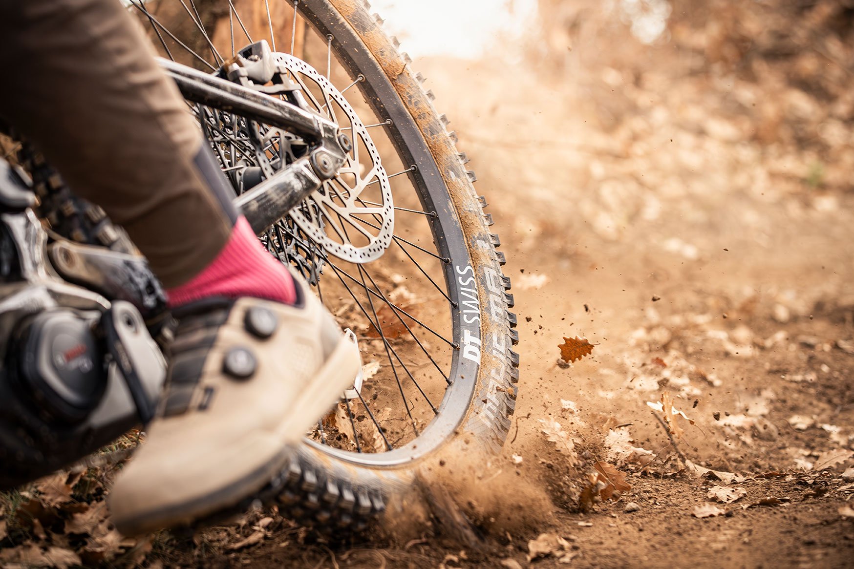Dynamische Nahaufnahme des Hinterrads eines Focus SAM² 6.9 E-Mountainbikes beim Driften auf einem Waldweg. Das DT Swiss Laufrad wirbelt trockene Blätter und Erde auf. Im Vordergrund ist der Fuß des Fahrers in einem beigefarbenen Klickschuh mit BOA-Verschluss und auffälligen pinkfarbenen Socken auf der Pedale zu sehen. Der Fokus liegt scharf auf der Bremsscheibe und dem Reifenprofil.